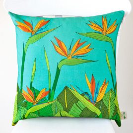 A handmade cushion featuring illustrated tropical bird of paradise flowers in oranges and blue with lots of green leaves set against a deep turquoise background, shown on a white chair