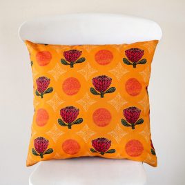 A handmade cushion featuring lino cut style proteas, vintage doilies and African patterning on an orange background, shown on a white chair