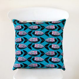 A handmade cushion featuring rows of lino cut style fish in black, with colourful scales in pinks, oranges, blues and white on a teal background, shown on a white chair