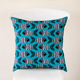 A handmade cushion featuring rows of lino cut style fish in black, with African-inspired patterning in pinks, oranges, blues and white on a teal background, shown on a white chair